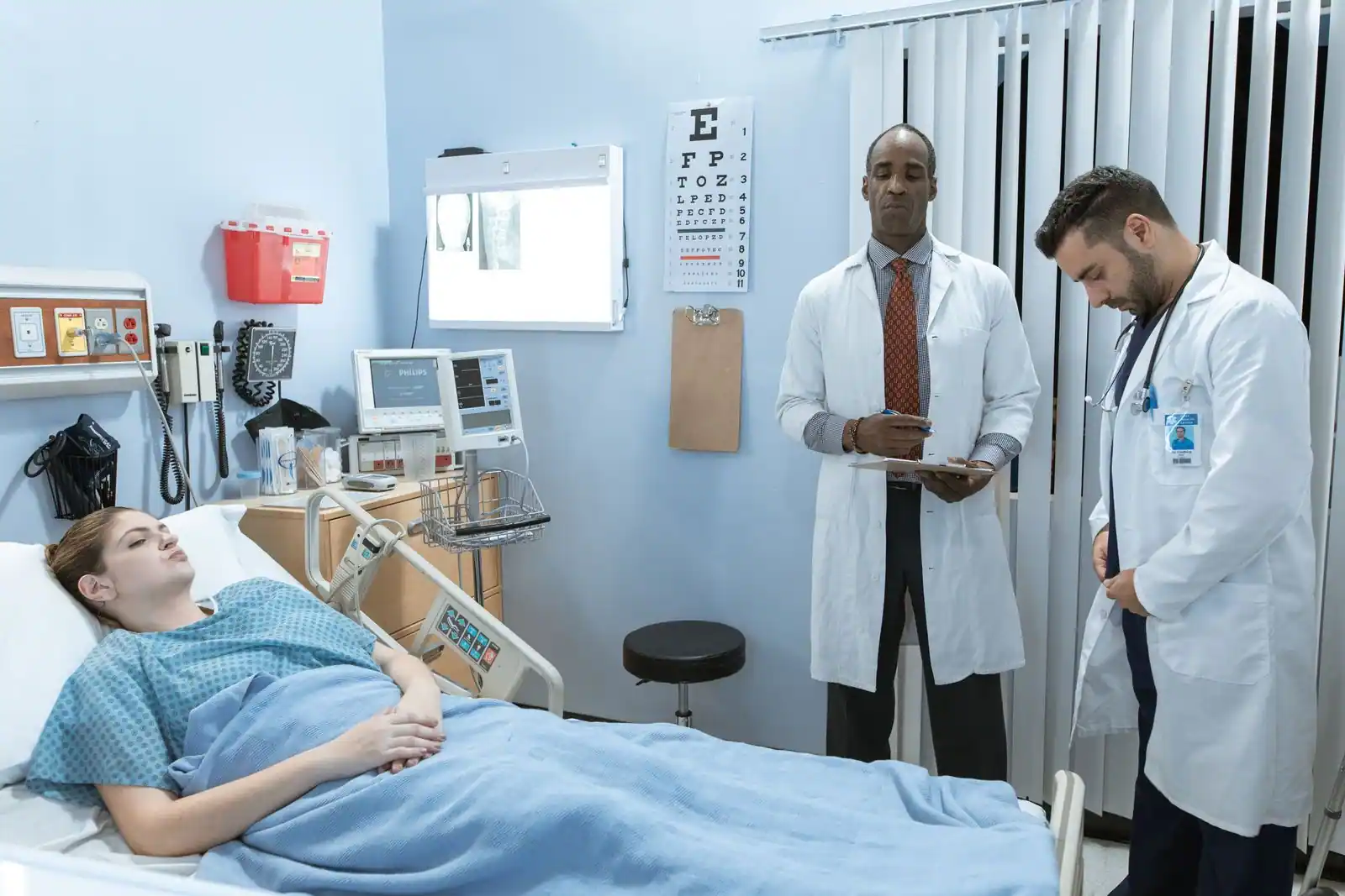 Patiente allongée dans un lit d'hôpital, deux soignants à son chevet entourés de matériel médical.