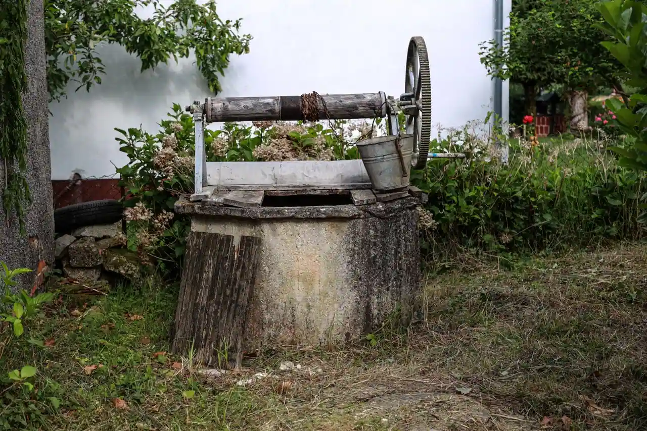 Puits busé dans un jardin, avec sa cuve en béton, son treuil à manivelle et un seau métallique