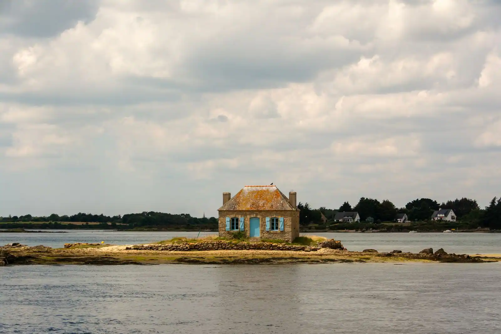Petite maison traditionnelle en pierre aux volets bleus sur un îlot, au milieu d'une ria de Bretagne entourée d'eau