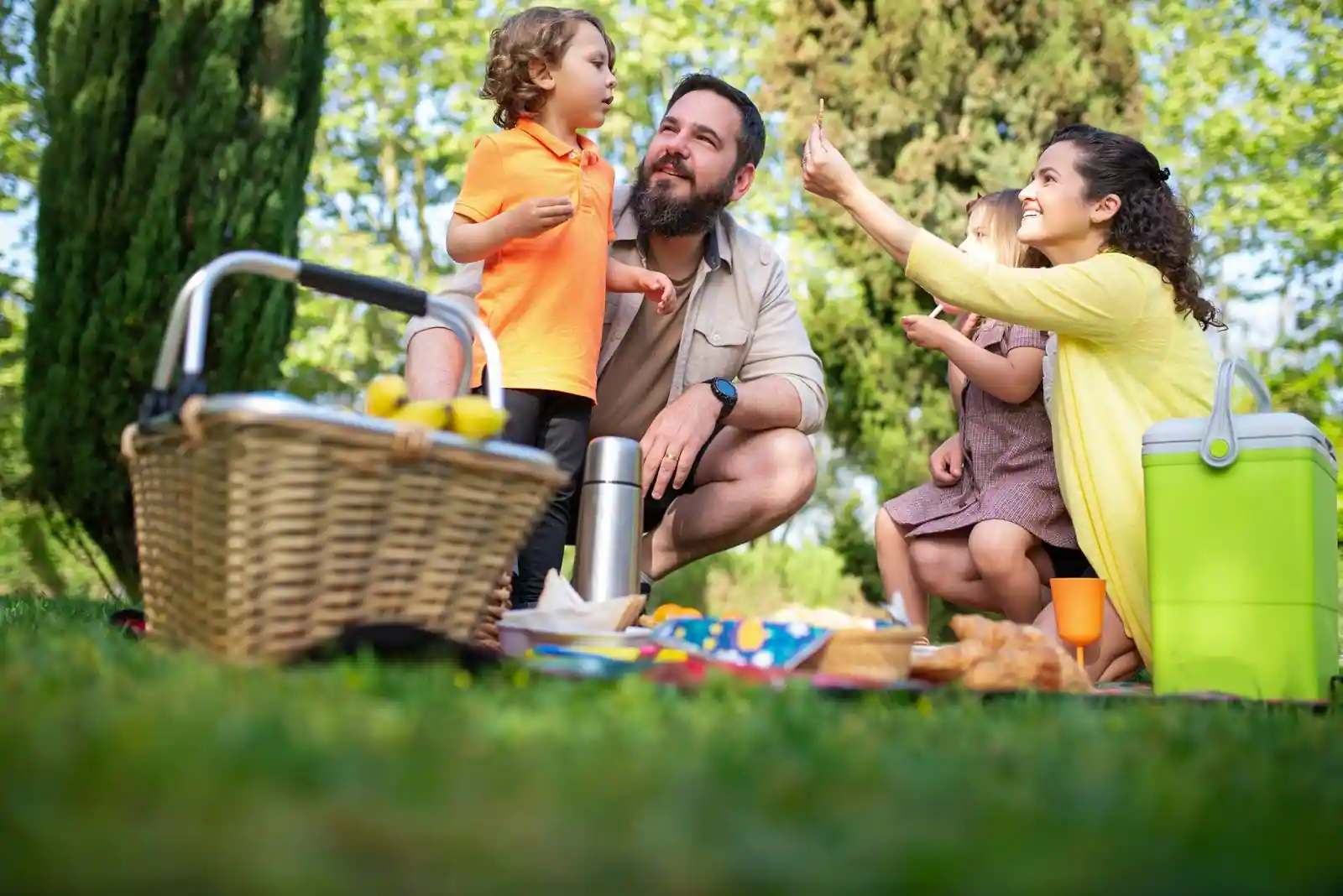 Famille avec enfants en train de pique-niquer sur l'herbe d'un parc, panier, glacière et en-cas posés devant eux