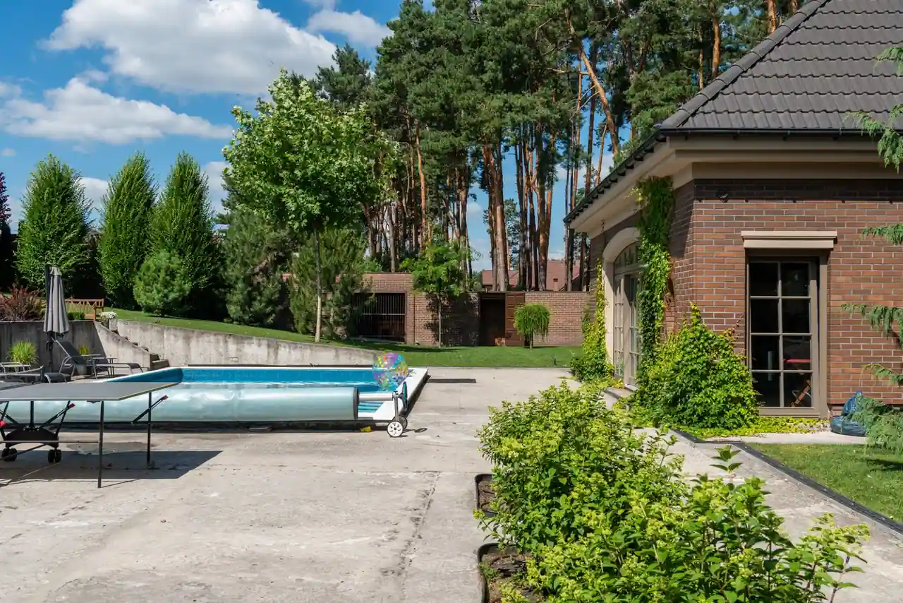 Piscine enterrée au bord d'une terrasse, à côté d'une maison en brique et d'un jardin arboré