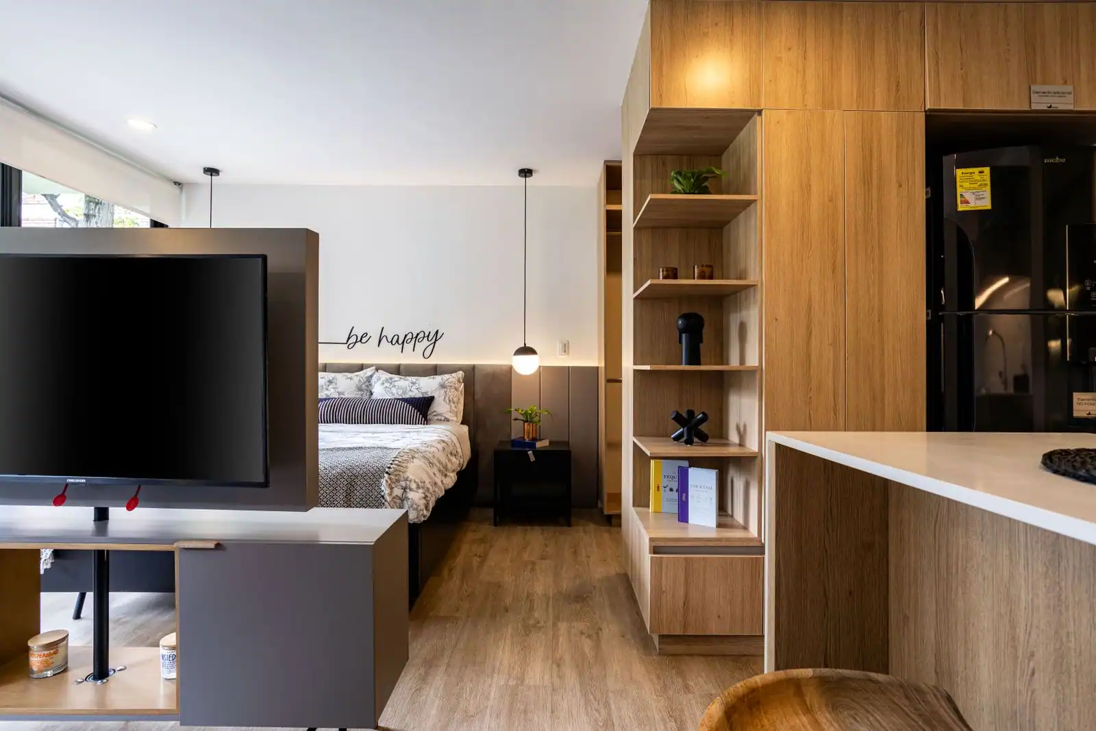 Intérieur d’un studio meublé moderne : lit, téléviseur, étagères en bois et kitchenette dans une seule pièce.
